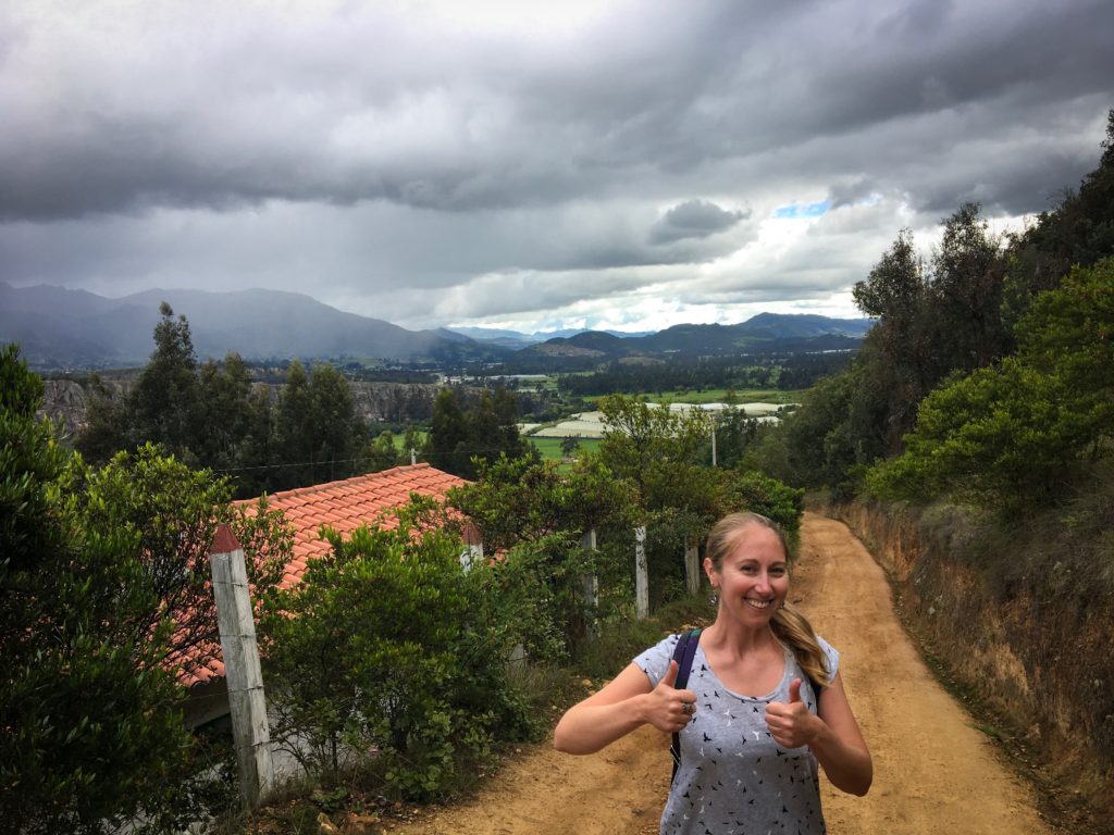 Alyssa giving a thumbs up to the camera on a hike. Mountains in the background.