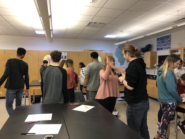 A classroom of students are walking around interacting with each other. Two students in the front are High-fiving.