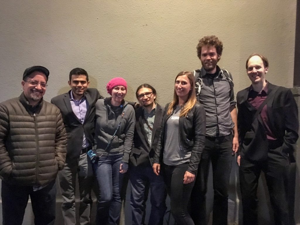 Seven people standing against a gray wall, smiling.