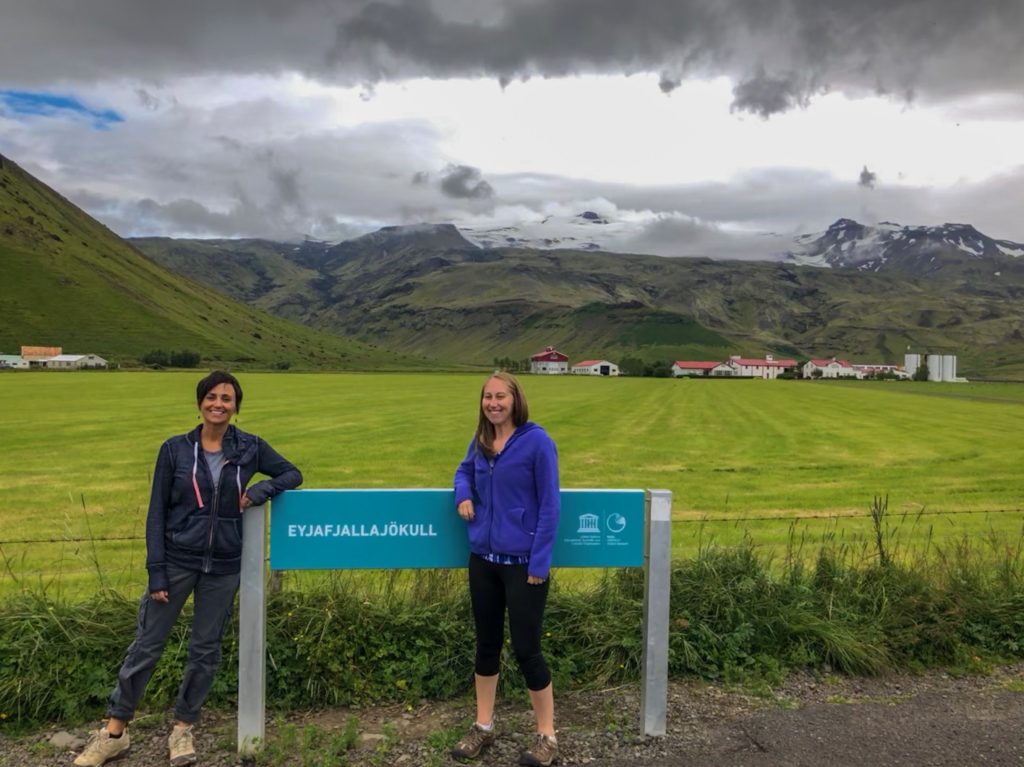 Two woman leaning on a sign for Eyjafjallajokull Volcano with the volcano in the background.