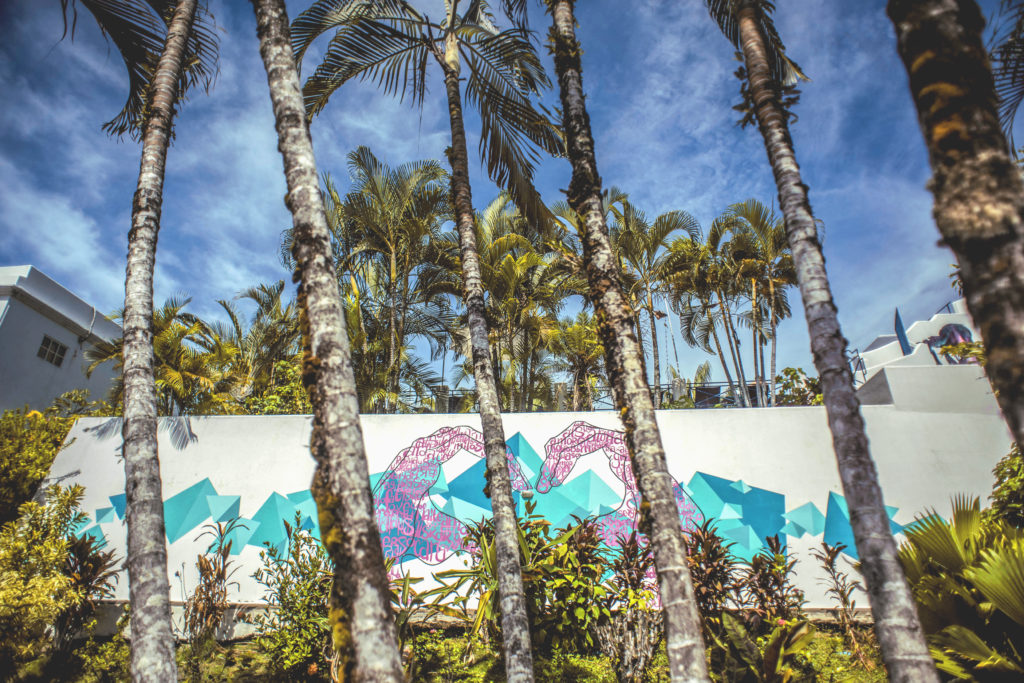 Palm trees in front of white wall. Mural painted on wall shows pink hands forming a heart at Selina Hostel