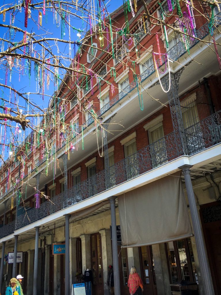 Mardi Gras beads hanging from tree in front of traditional New Orleans building.