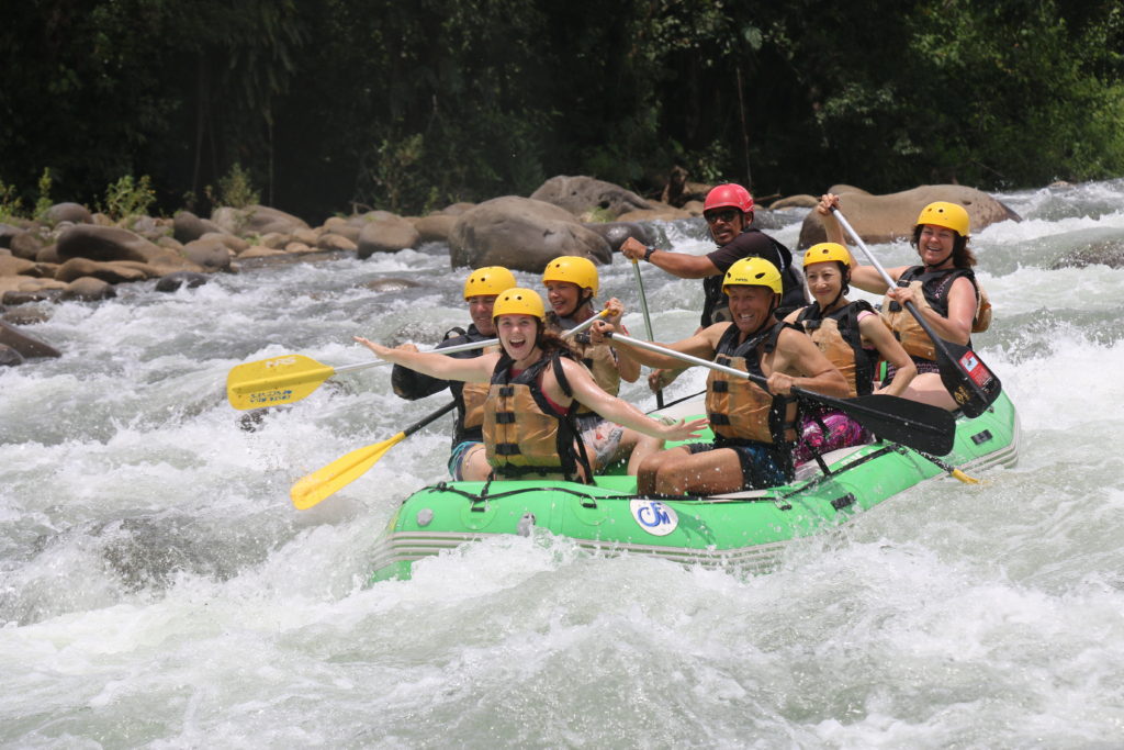 Seven people in an inflatable raft white water rafting. Trees in the background.