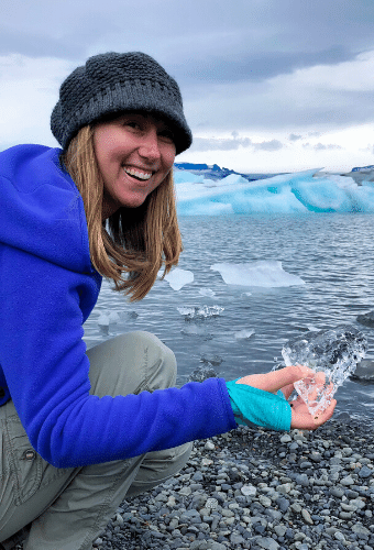 Alyssa kneeling down holding a piece of ice from a glacier in front of an ice lagoon.