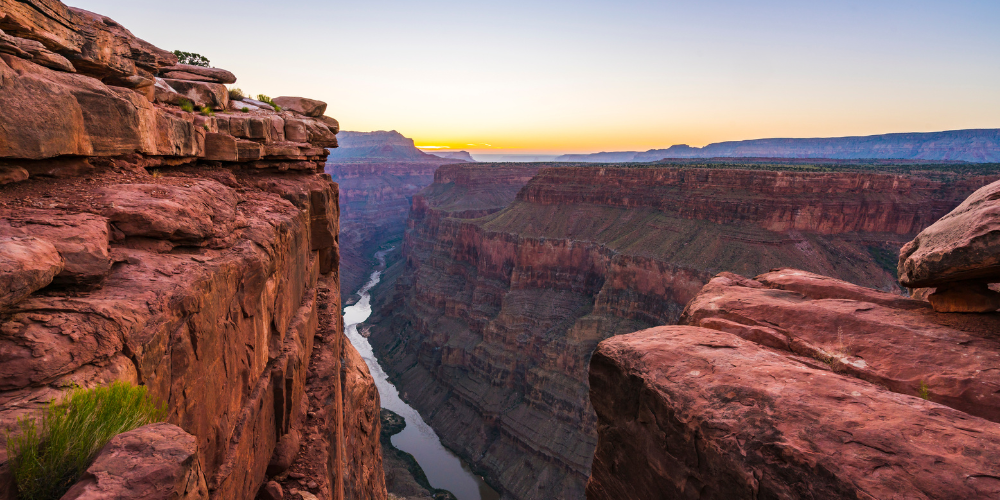 Colorado River cuts through the red layered rock of the Grand Canyon as the sun sets in the background.